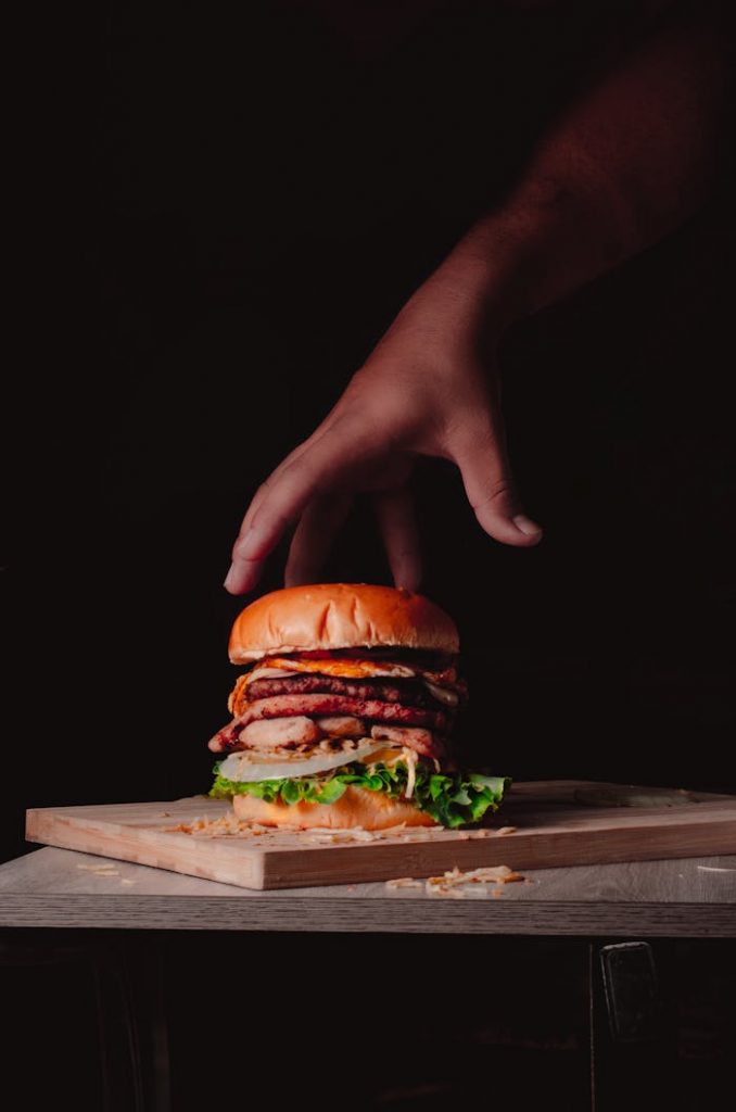 Close-up of a hand reaching for a gourmet burger with various toppings on a wooden board.